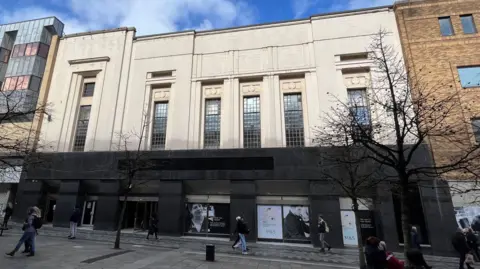 A former retail building in darkness. There are some old M&S posters in the windows. A few shoppers are walking past. There are some small tree in the foreground. 