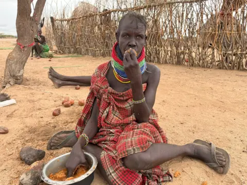 A woman with a cloth tied round her body sits on the dusty ground. She has her hand in a bowl of fruit.