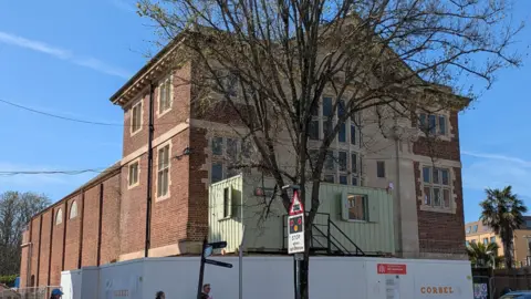 Paignton Picture House Trust The exterior of Paignton Picture House on a bright blue sky day. There is fencing around the front and a container with two windows is parked above it. In front of the building is a tree and a sign indicating a level crossing. There is a palm tree to the right of the building.
