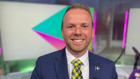BBC Ben Maguire is smiling broadly at the camera. He is wearing a white shirt, blue jacket and a Cornish tartan tie. He has short blond hair and a slight beard. There is St Piran flag badge in the button hole of his jacket. Behind him are neon-lit panels.