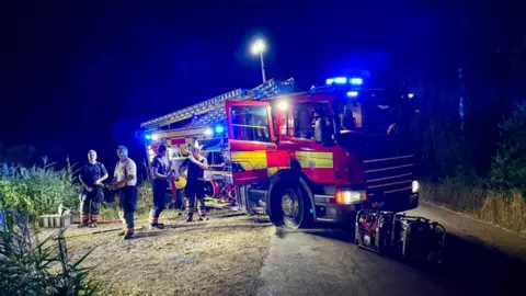 Peterborough Volunteer Fire Brigade A night-time image showing four men by their fire engine, which is lit up with artificial lighting. There is greenery all around.