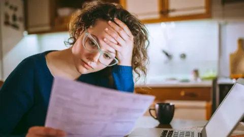 A woman holds her forehead in stress while reading a form in front of her laptop.