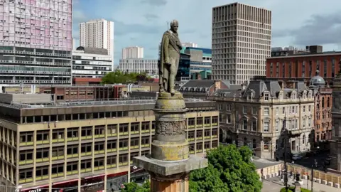 A statue of Sir Walter Scott high on a pillar. Greenery and buildings are in the background.