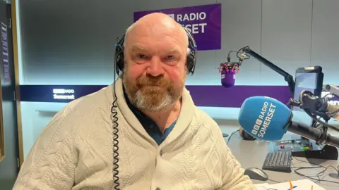 Council leader Bill Revans wears a cream coloured knitted cardigan. He sits at a desk in the BBC Radio Somerset Studio and has a pair of headphones on his head