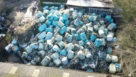 An aerial view of bales of rubbish which have been dumped on a farm in Lincolnshire. The blue-coloured bundles are piled up and surrounded by two agricultural buildings.