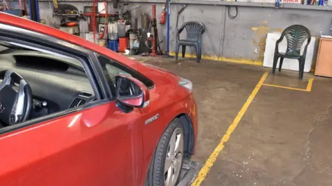 A red car within yellow lines on the floor in a garage. There is a dark green plastic chair in front of the car, slightly to the right, within separate yellow lines on the floor. 