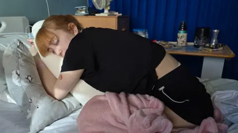 Millie Fitzsimons Woman with ginger hair on a hospital bed crouched over a pillow.
