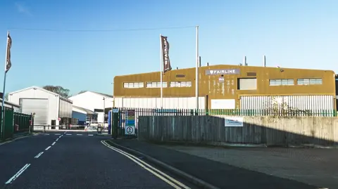 Ant Saddington/ BBC A yellow and white corrugated iron building surrounded by a green metal fence. There is a road in the foreground, with double yellow lines painted on it.