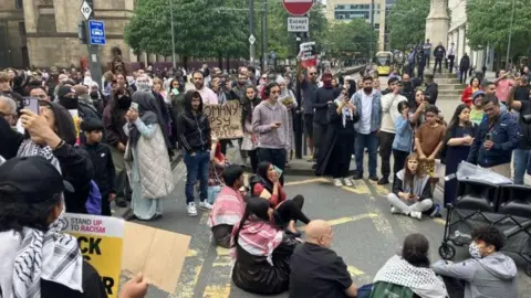 BBC/Phil McCann Proetsters gather in Manchester - several are seated on the floor with a yellow tram in the background