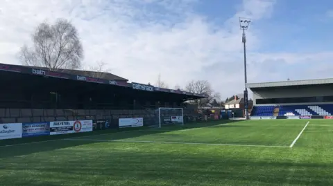 Two football stands adjacent to a grass pitch, one is a standing room only terrace and the other has blue seats with areas of white seating in the shape of an Adidas logo. 