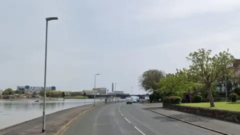 Google Maps image of the road running along Walney Island's Promenade. Several vehicles are travelling along the road, which has water on one side and grass and trees on the other.