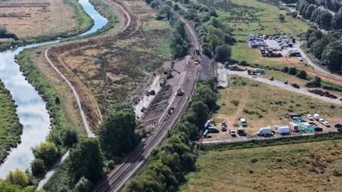 Eddie Mitchell An aerial view of a trainline running through countryside, with a river running almost parallel on the left-hand side and a town in the distance on the far right.
There are vehicles and diggers around the embankment in the centre of the image.