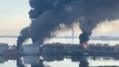 Volodymyr Zelensky/X Phone footage of black smoke rising above the St Petersburg oil terminal, three parts of the facility appear to have been hit. 