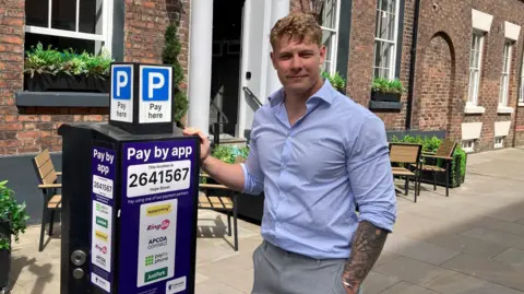 Carl Cashman has short blonde hair and is standing on a street next to a parking pay machine. He is wearing a light blue shirt and is looking at the camera.