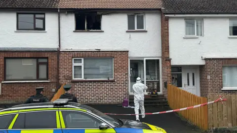 A terraced house between two others. An upstairs window is brunt out. The scene is cordoned off by police. Two officers are standing in front of a police car while another in white forensic suit is heading towards the front door of the house.