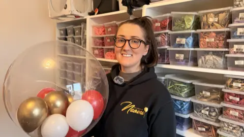 A lady in a black jumper, she has glasses on with her hair in a high pony tail. She's smiling at the camera stood in front of shelves with balloons in. The lady is holding a large balloon she had just made for a customer. 