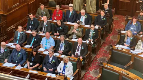 A bird's eye view of a council chamber, focused on several rows of seats featuring people wearing turquoise rosettes, identifying them Reform UK councillors