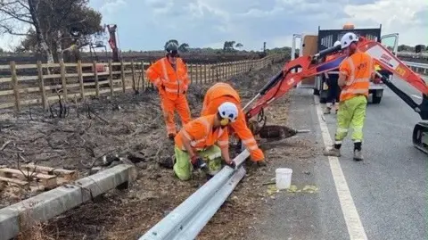 Four people work on a metal safety barrier at the side of a road. They are wearing orange and yellow high-viz clothing. The land to the side of the road is charred black.