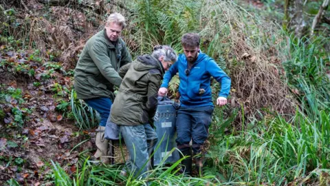 Sam Rose Three people carrying a large cage down a steep grassy bank. They are wearing blue jeans. Two are wearing Barbour-type jackets and the other is wearing a Royal blue fleece.