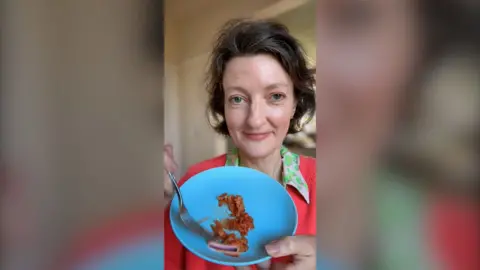 A woman with brown hair holds up a blue plate holding kimchi. She is wearing a dark pink jumper and a green-collared shirt.
