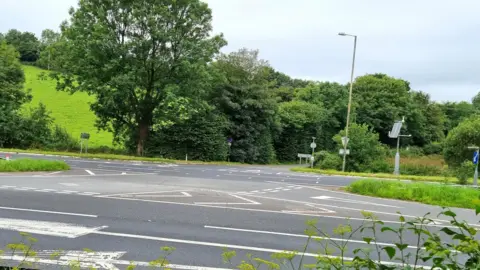A road junction on a dual carriageway. There are green trees in the background. The sky is grey. 