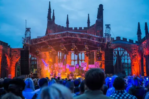Getty Images The ruins of Coventry Cathedral can be seen behind a stage which it lit in reds and oranges with the sky blue above. The Specials can be seen on stage with members of the crowd in the forefront. 
