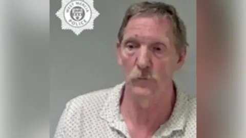 Mug shot of a white man with short hair and a moustache. He is wearing a patterned white polo shirt. 