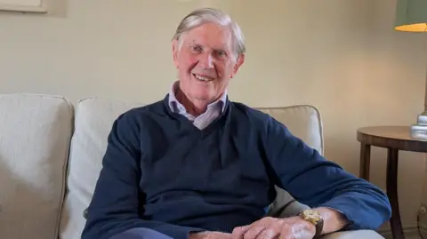 Bill Cash wears a navy V-neck jumper over an open button collar shirt. He is smiling at the camera. He is sat on a sofa next to a table with a lamp on it. 