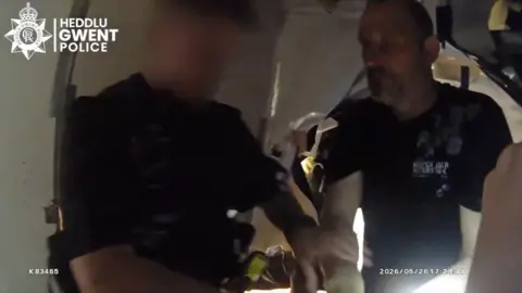 This image shows body cam footage of a man being arrested, he's in a dimly lit room and dressed in a black t shirt..