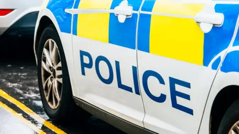 A car with Police written on the side is parked beside double yellow lines on a wet street