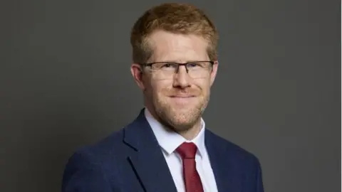 Thomas Collins/House of Commons/Laurie Noble A man with ginger hair wearing glasses, a blue suit, white shirt and red tie smiles at the camera