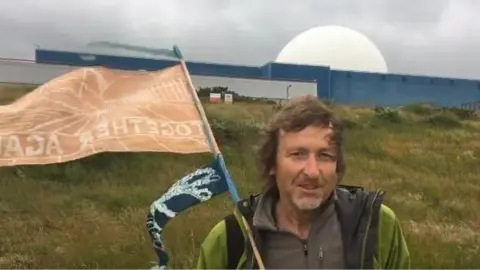 Contributed A man stands in front of a large power station on a grass bank. He has medium length brown hair and is wearing a green coat and brown jumper. He is holding an orange flag.