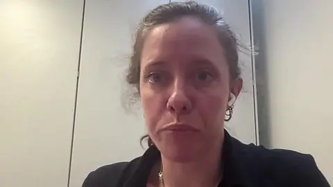 A woman in her 30s with light brown hair tied back and a black jacket speaks into a camera