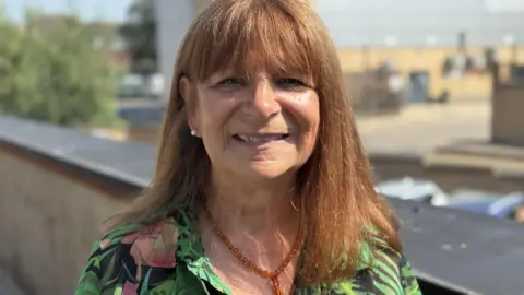 A woman wearing a green top is smiling for the camera. She is wearing a brown translucent necklace and white earring. There is a wall and buildings behind her.