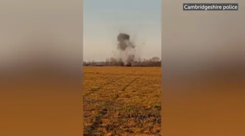 A plume of smoke rising at the other side of a field. Clods of earth are shooting into the sky.