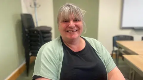 A woman with blonde hair tied up smiles at the camera. She is sat in a chair, with a black t-shirt and green shirt over the top.