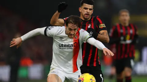 Reuters Luton Town's Tom Lockyer in action with AFC Bournemouth's Dominic Solanke. Lockyer is wearing a white Luton Town football kit with an orange vertical stripe down the right hand side and has a long-sleeved white undershirt on. He is sticking his tongue out in concentration as he battles for the ball. Behind him, Solanke is wearing a red and black striped Bournemouth shirt with black shorts and black gloves. He has his left hand on Lockyer's shoulder as he tries to get the ball off him. The yellow ball can be seen in front of Lockyer.