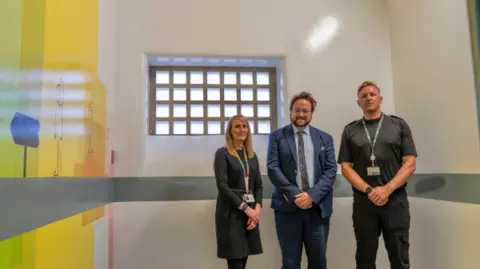 Cleveland Police Three people stand in a small white room facing the camera. The new "youth focussed custody suite" has a colourful painting on one wall. The small window at the top of the room has a criss-crossed bar patterned frame.