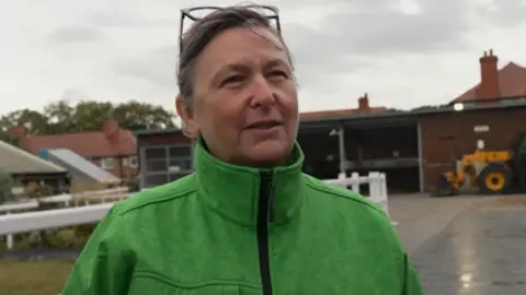 A woman wearing a lime green rain coat and with glasses on top of her head. She is smiling as she is interviewed, standing in the rain in a racecourse stable yard.
