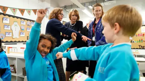 PA Media Elke Budenbender pictured in a primary school. She stands in between two women, one of whom is pointing towards something off to the right. In front of them stand two small children in turquoise school uniform, including a little girl smiling at the camera and holding her arms above her head.