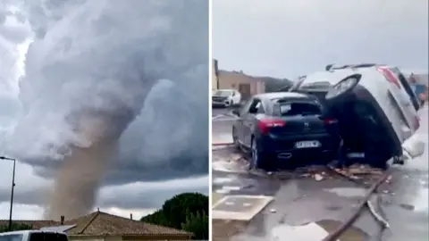 A tornado is seen spinning above a house in the clouds, with two cars on top of each other on the right.