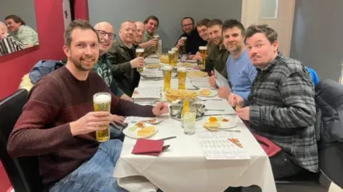 Dean Sullivan The ten friends sit round three sides of a table in a curry house and pose for a picture. Some are holding pints with other drinks on the table along with menus.