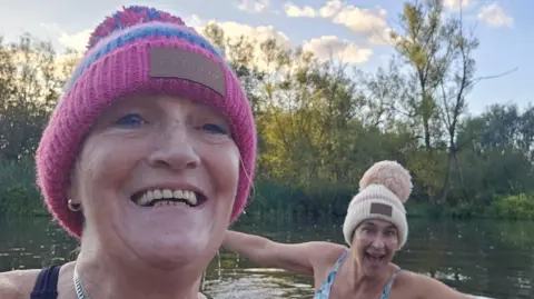 Lisa Thomas Two ladies in a river both with bobble hats on, one pink the other beige, one has a floral blue swimsuit, the other a black one, behind them are trees.