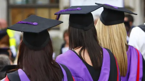 A file photo of university graduates. They are standing with their back to the camera and are wearing black robes with purple hoods and black graduation caps. All three have long straight hair.
