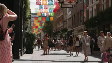 A busy street in central London with colourful banners and people in summer clothes 