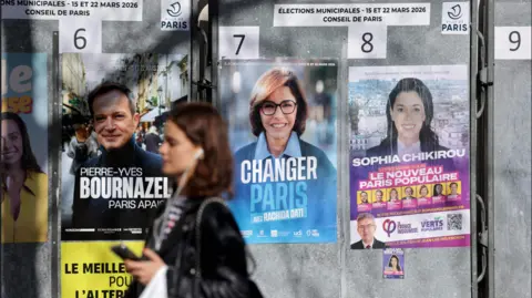Getty Images A woman walks past campaign posters of Paris mayoral candidates. The posters visible are of Sophia Chikirou, Rachida Dati and Pierre-Yves Bournazel