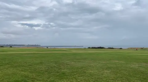 The Leas on the Coast Road at South Shields. There is grass in the foreground and sea in the background with stormy skies above.