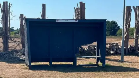 Jersey Wanderers Football Club A dark blue sports team shelter stands on a dry grass field. The shelter has a damaged side panel with a large hole. Cut tree trunks, stacked logs, and a lamp post are visible in the background under a clear blue sky.