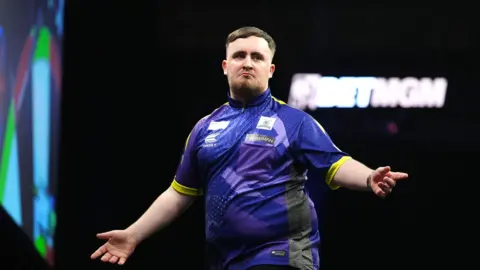 PA Media Luke Littler playing darts. He is looking off camera and has his arms stretched out wide. He is wearing a purple coloured darts top. 