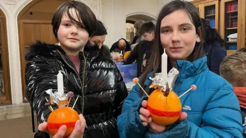 Violet and Scarlett hold the Christingles they have made in the vestry at Great Yarmouth Minster. Violet is wearing a black jacket and Scarlett is wearing a blue coat. Behind them are other children and against the wall is a row of fitted bookshelves.